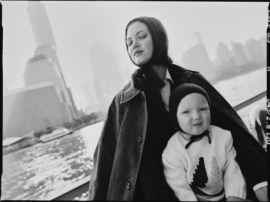 Black and white photograph captures a mother and child outdoors with a city skyline and waterfront visible in the background.