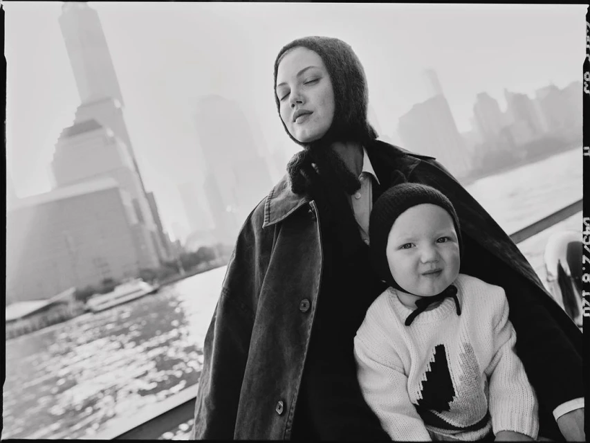 Black and white photograph captures a mother and child outdoors with a city skyline and waterfront visible in the background.