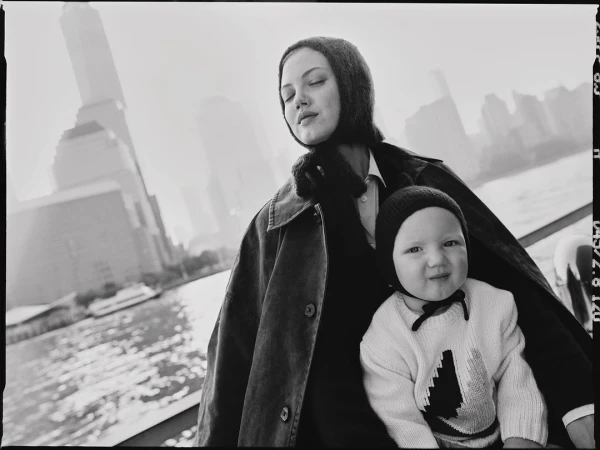 Black and white photograph captures a mother and child outdoors with a city skyline and waterfront visible in the background.