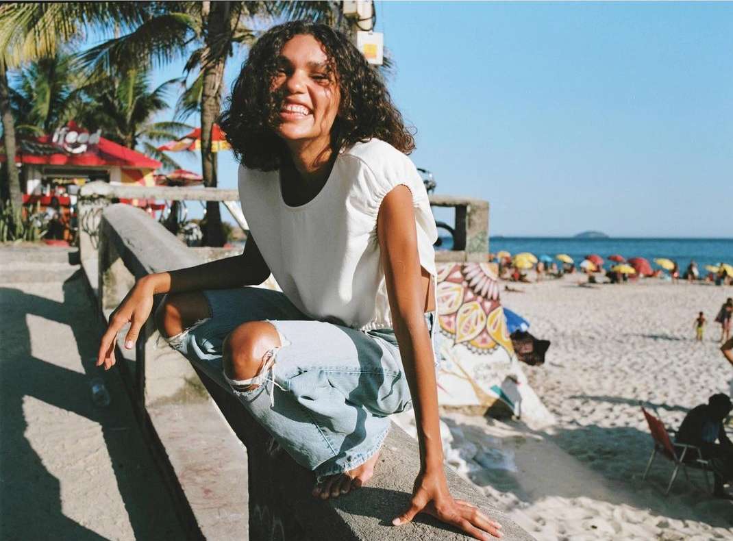 Smiling young woman with curly hair sits on a concrete ledge overlooking a sunny beach with parasols and palm trees in the background.