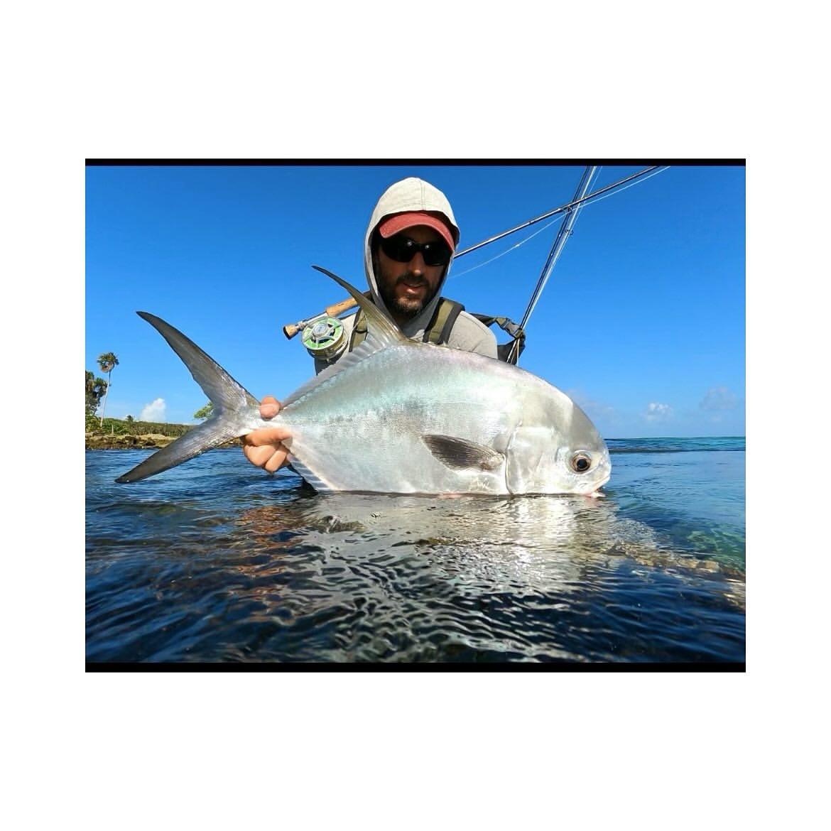 Fisherman displays a large silvery fish while wading in clear water on a sunny day, showcasing a successful catch and scenic view.