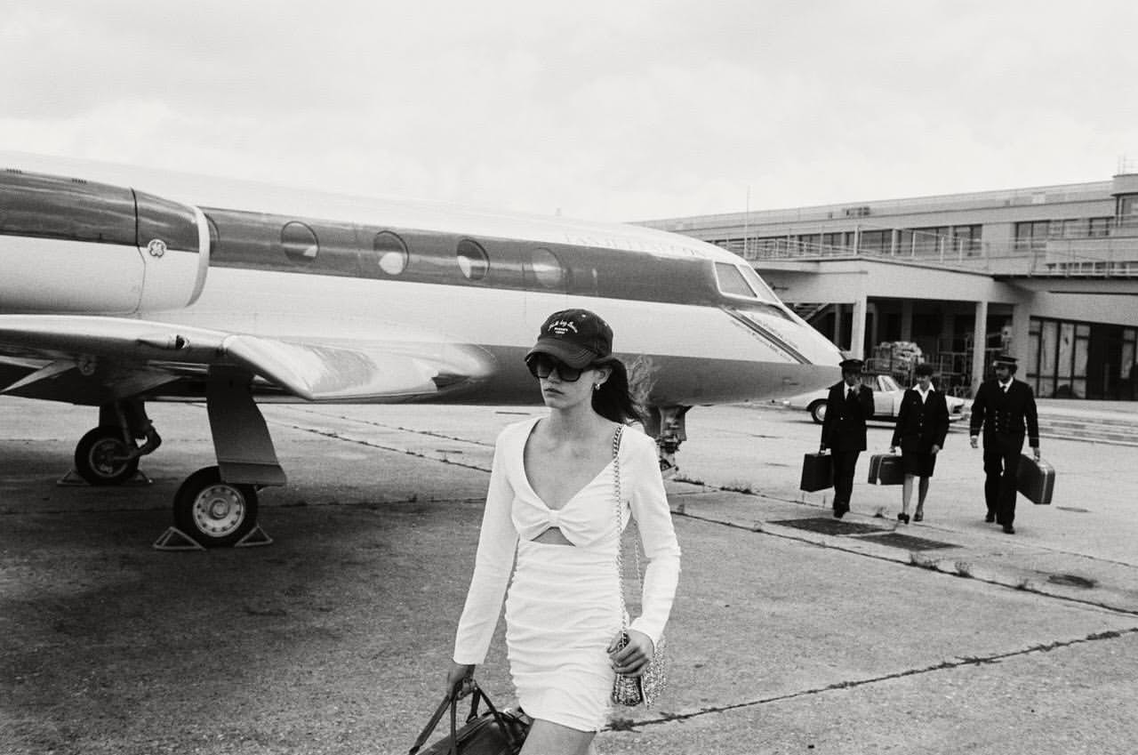 Black and white image of a woman in a white dress walking in front of a private jet, with pilots walking behind.