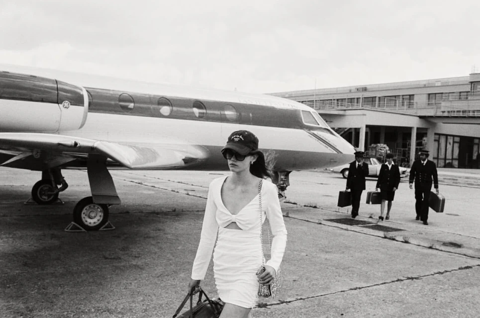 Black and white image of a woman in a white dress walking in front of a private jet, with pilots walking behind.