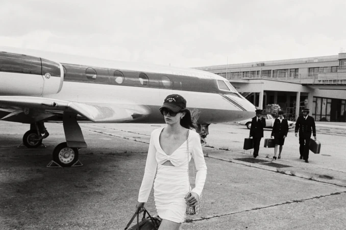 Black and white image of a woman in a white dress walking in front of a private jet, with pilots walking behind.