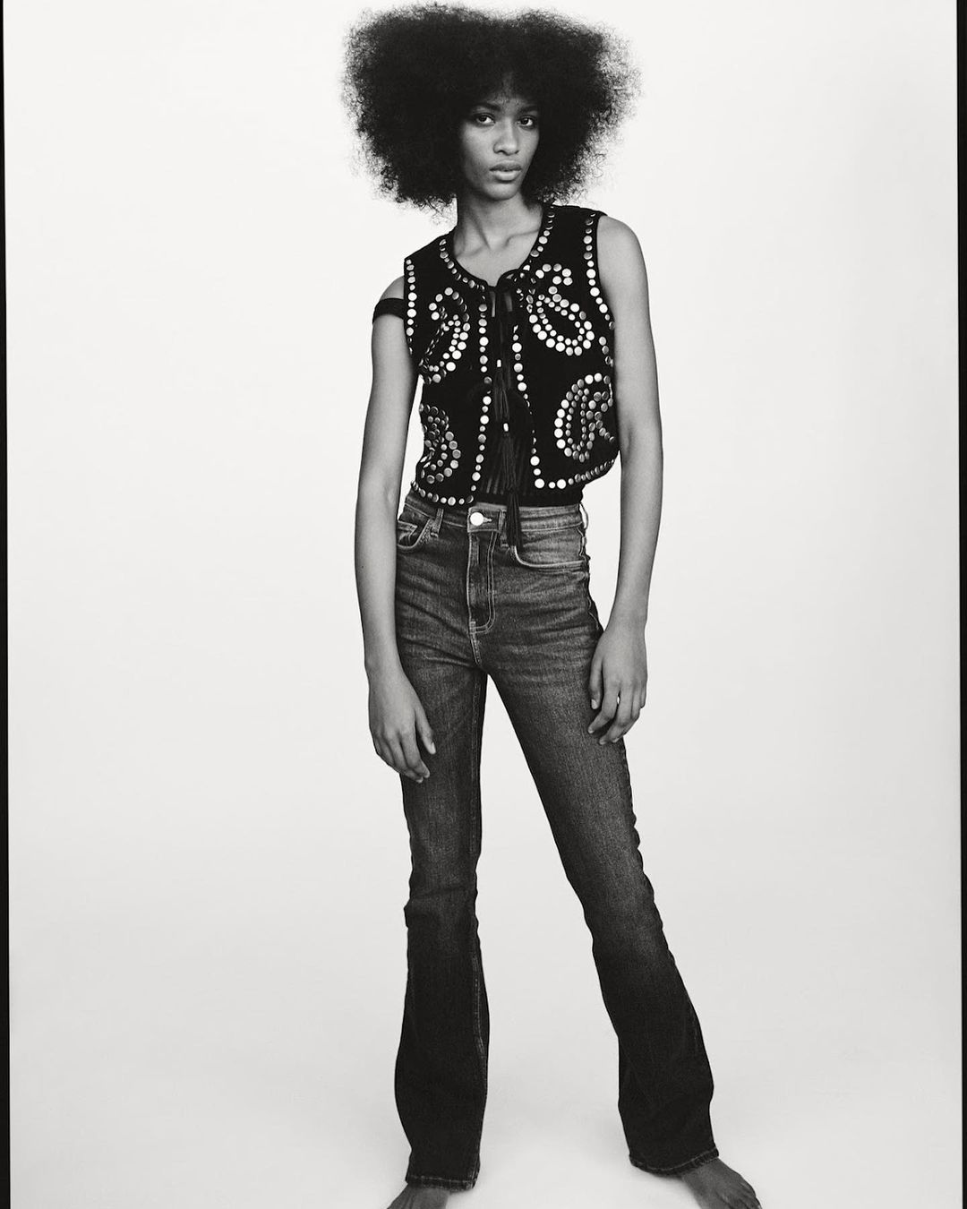 Monochrome portrait of a Black female model with afro, wearing a studded vest and jeans, posing against a white studio background.