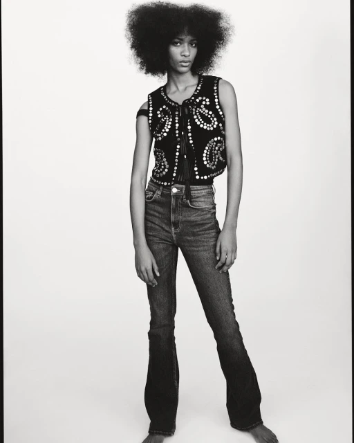 Monochrome portrait of a Black female model with afro, wearing a studded vest and jeans, posing against a white studio background.