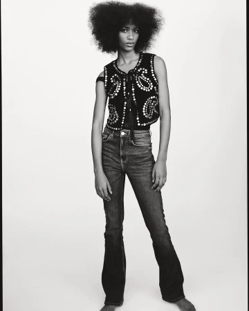 Monochrome portrait of a Black female model with afro, wearing a studded vest and jeans, posing against a white studio background.