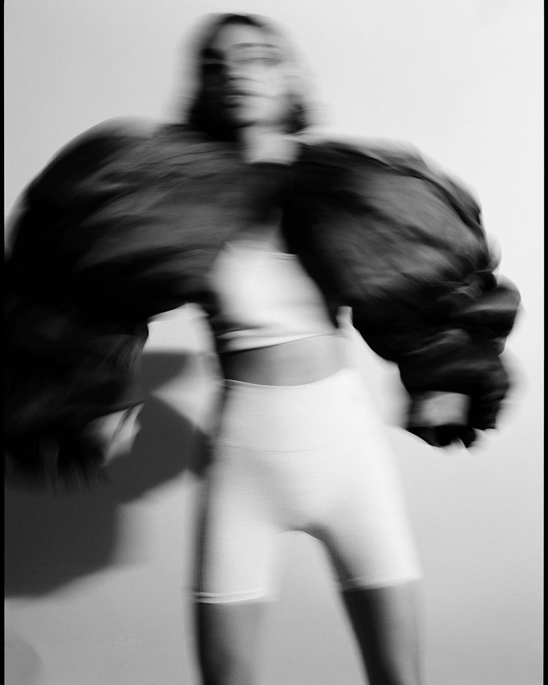 Blurred fashion portrait of a slender adult woman posing in a white crop top and biker shorts with an oversized jacket in grayscale.