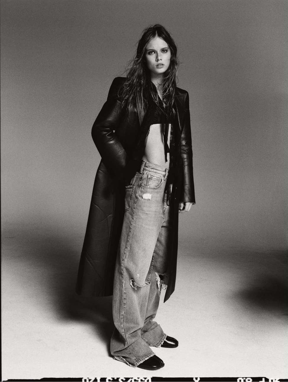 Monochrome portrait of a stylish woman in a black coat and ripped jeans. She poses against a gray backdrop in a studio.