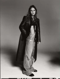 Monochrome fashion portrait of a model wearing a dark leather coat and distressed baggy jeans against a plain studio background.