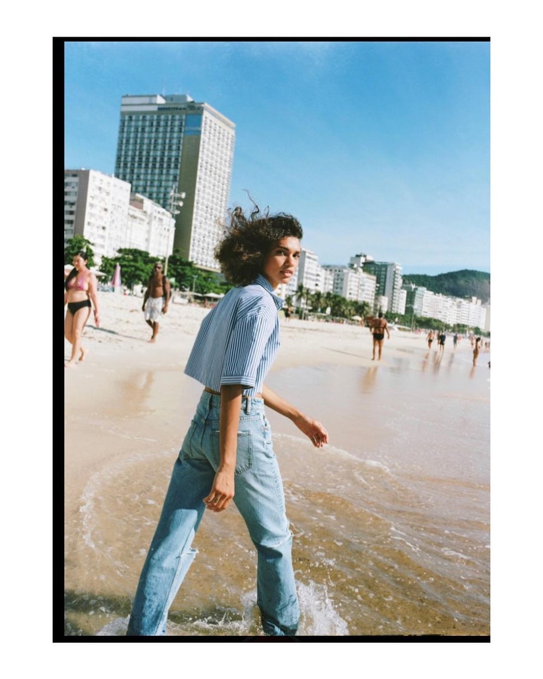 Woman walking on sandy beach wearing blue striped shirt and denim jeans with urban backdrop
