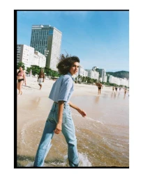 Woman walking on sandy beach wearing blue striped shirt and denim jeans with urban backdrop