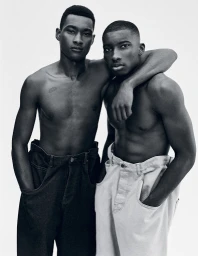 Monochrome portrait of two shirtless men standing together with one arm around the other against a white background.