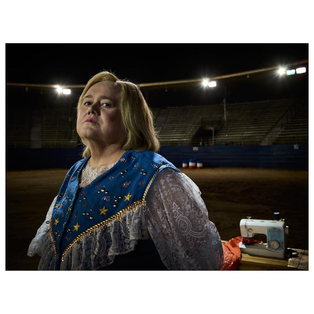 A woman in a blue and gold vest stands near a sewing machine at night in front of a stadium-style seating structure outdoors.