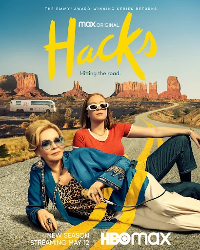Two adult women lying on a highway with desert landscape in the background. One woman is wearing sunglasses, blue top and leopard print top.