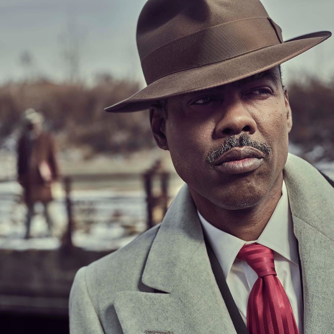 Man wearing brown fedora and grey overcoat with a red tie stands outdoors with a blurred figure in the background.