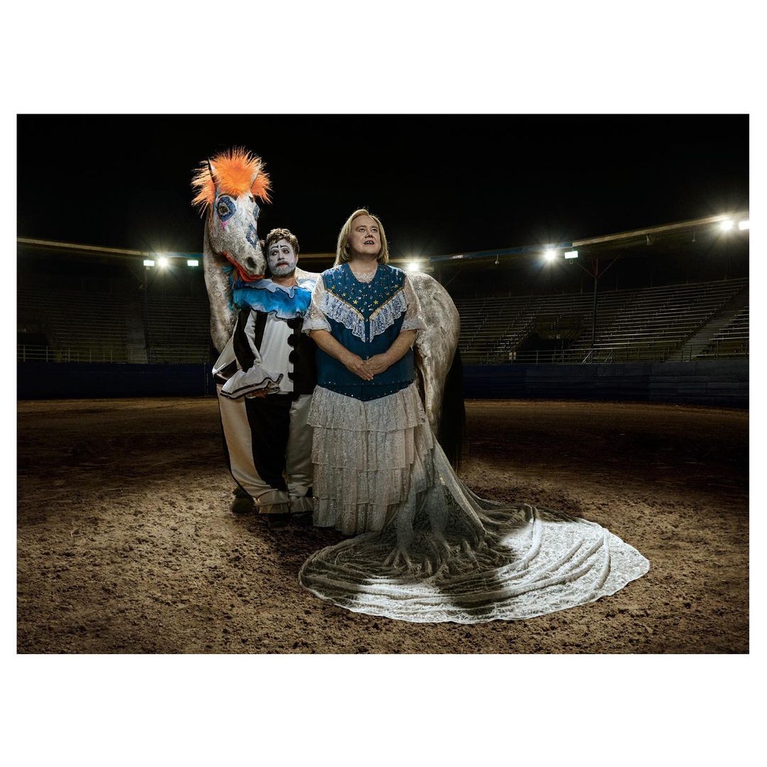 Rodeo arena scene with a woman in a long dress, a man in clown makeup, and a horse with an orange mane performing in a spotlight.