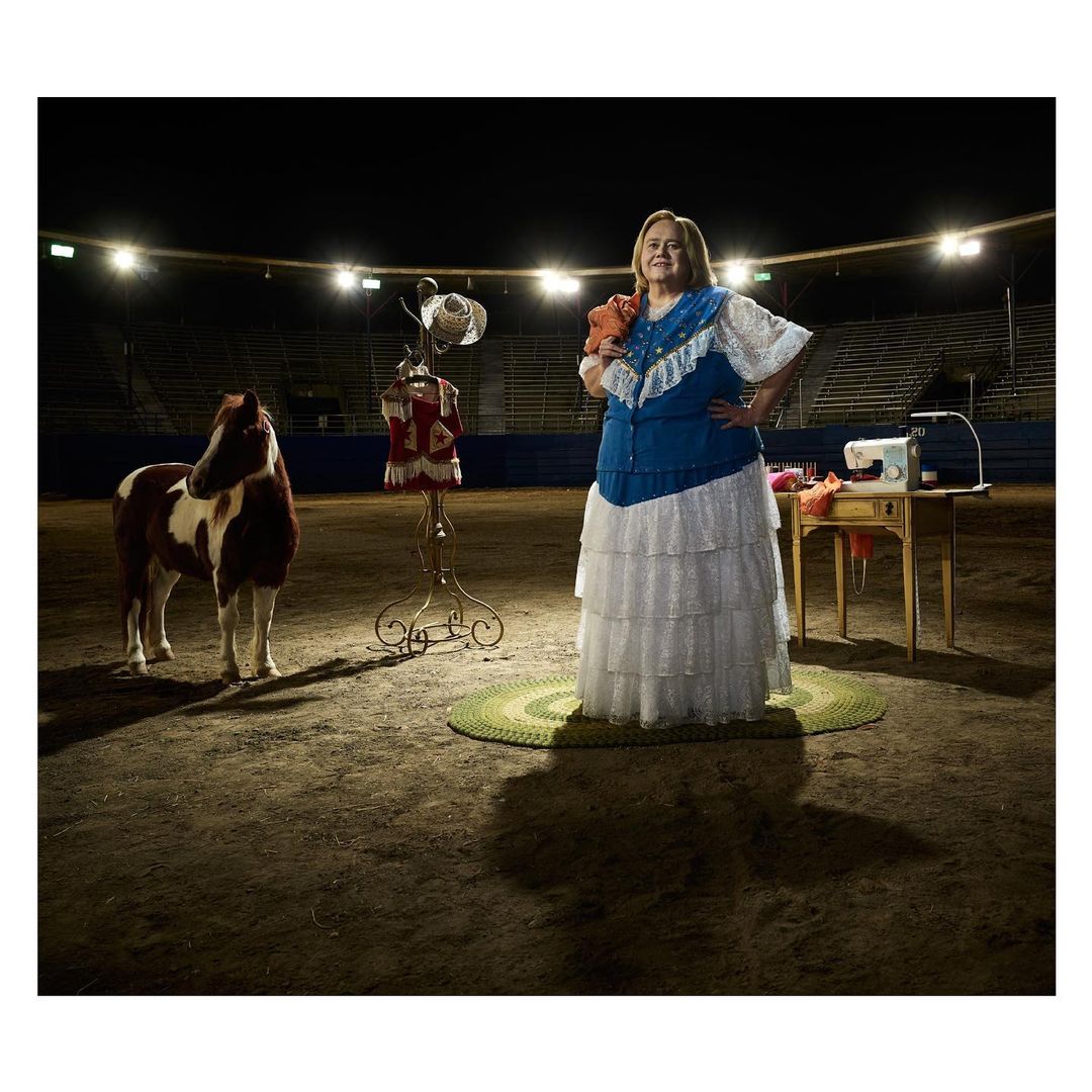 Woman in costume stands next to a pony, sewing machine, and dress form inside an empty stadium, bathed in bright overhead lighting.