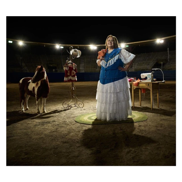 Woman in costume stands next to a pony, sewing machine, and dress form inside an empty stadium, bathed in bright overhead lighting.