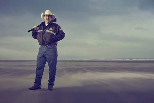 Man wearing a white cowboy hat, dark jacket, and blue jeans standing on a sandy beach holding a rifle across his shoulders.