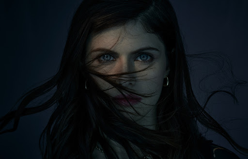 Close-up portrait of actress Alexandra Daddario with dark hair blowing across her face and striking blue eyes against a dark background.