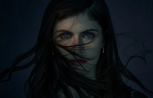 Close-up portrait of actress Alexandra Daddario with dark hair blowing across her face and striking blue eyes against a dark background.