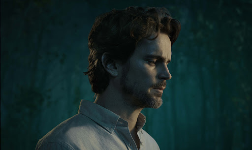Close-up studio portrait of a male with dark hair and a light shirt, set against a blurred teal background.