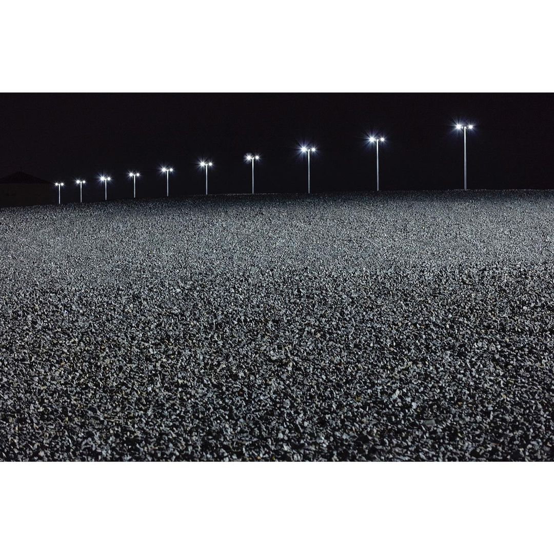 Wide outdoor shot at night features row of lights illuminating dark asphalt road, creating a stark contrast in a dark, desolate environment.