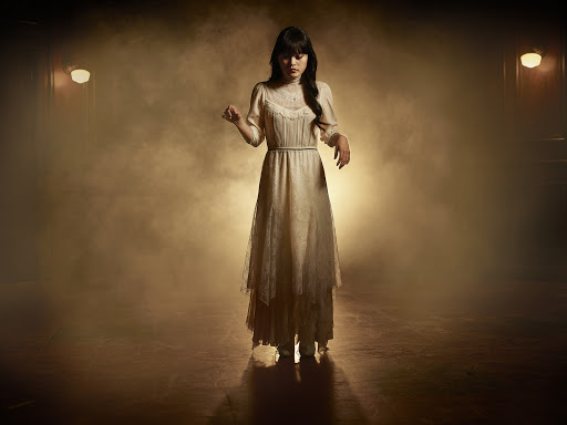 Woman in a vintage cream-colored gown stands in a smoky, theatrical setting, softly lit with visible light fixtures. Fashion, vintage style.