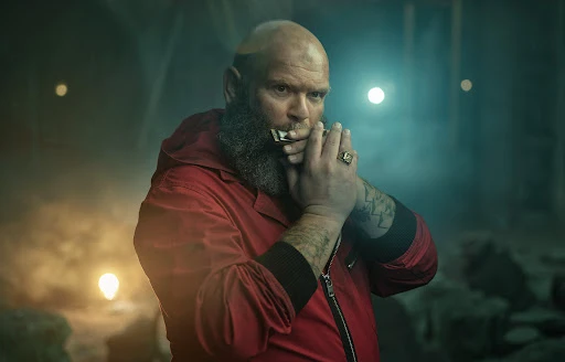 Close-up of a bald adult male with a beard wearing a red hooded jacket, holding a harmonica to his lips in a dimly lit indoor setting.