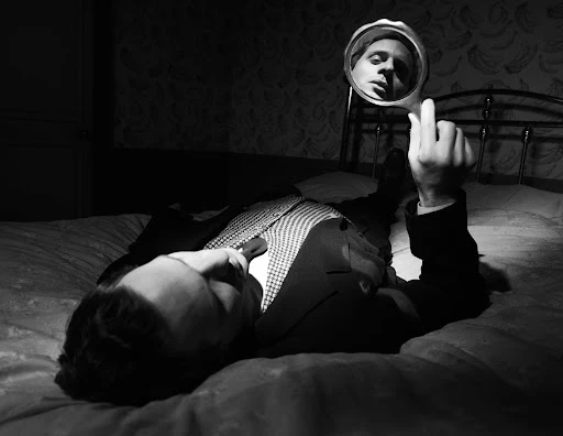 Black and white portrait of a man lying on a bed, holding a mirror reflecting his face in an indoor bedroom setting with wallpaper.