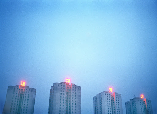 Four buildings with neon Chinese text on top against a light blue sky, captured at eye-level in an urban environment.