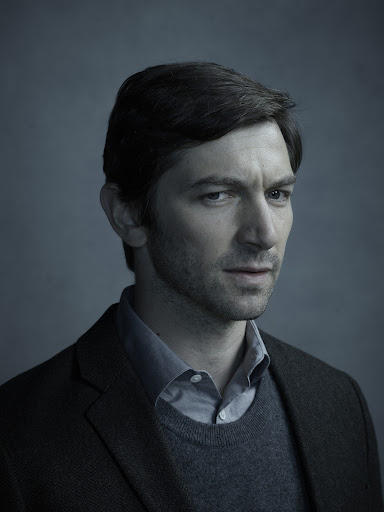Portrait of a man wearing a dark blazer over a grey sweater and collared shirt against a grey textured studio backdrop.