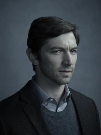 Portrait of a man wearing a dark blazer over a grey sweater and collared shirt against a grey textured studio backdrop.