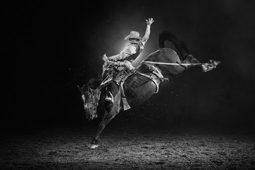 Black and white image of a cowboy riding a bucking bronco in a rodeo. The cowboy wears a hat and vest as the horse rears up.