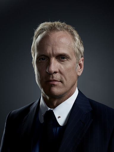 Headshot of a blond-haired adult male wearing a navy suit, collared shirt, and tie against a dark grey background, looking directly at the camera.
