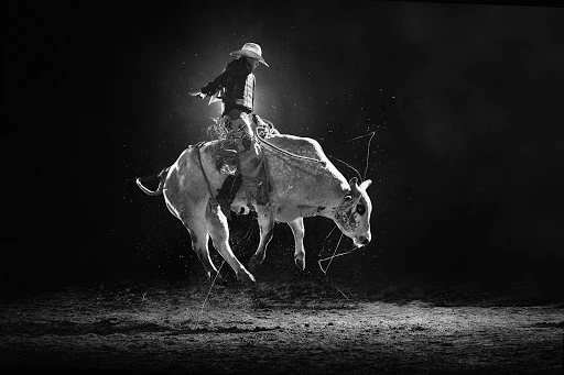 Monochrome shot of a bull rider wearing a cowboy hat on a bucking bull in a rodeo arena with a dark background.