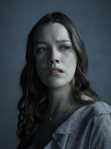 Close-up studio portrait of a Caucasian woman with long brown hair, wearing a patterned grey shirt, looking sad with tears on her face.