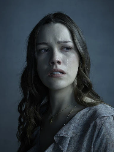 Close-up studio portrait of a Caucasian woman with long brown hair, wearing a patterned grey shirt, looking sad with tears on her face.