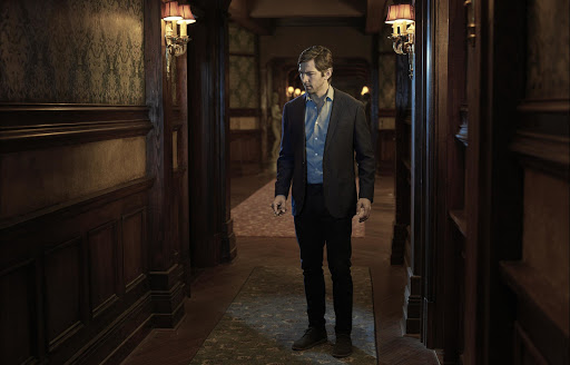 An adult male in a dark suit and blue shirt stands in a dim hallway with dark wood paneling and patterned wallpaper, lit by wall sconces.