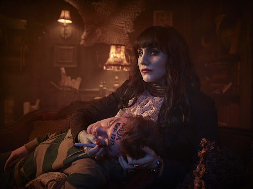 Woman with dark hair writing on a person's face with a marker in a dark indoor setting, lit by table lamps in an intimate moment.