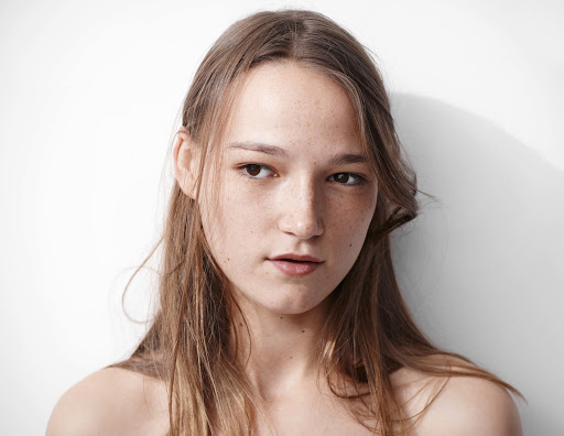 Close-up portrait of a young woman with freckles and long brown hair looking away against a plain white background