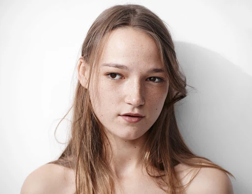 Close-up portrait of a young woman with freckles and long brown hair looking away against a plain white background