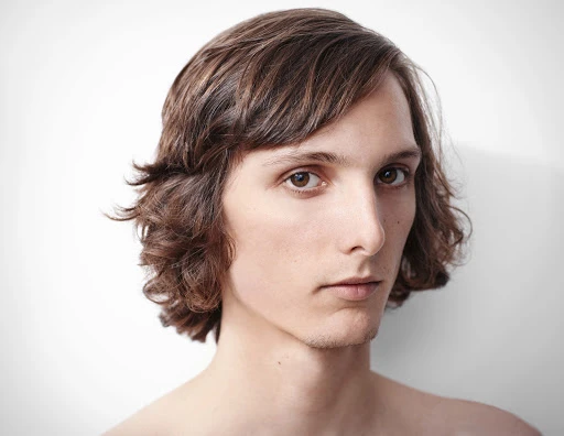 Portrait of a young adult male with brown wavy hair, light skin, and brown eyes, against a plain white studio backdrop, looking directly at camera.