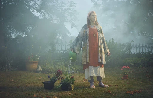 Elderly woman in garden watering plants with green watering can; misty outdoor scene with garden, plants, and flowers.