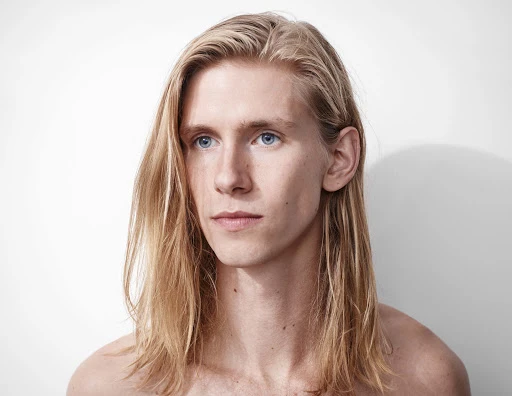 Portrait of a young man with long blonde hair and blue eyes against a plain white studio background.