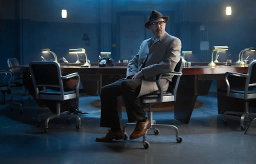 Man in hat and coat sits in an office setting with vintage phone, desk lamps, and rolling chair under blue lighting. 