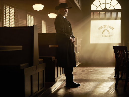 An older adult male with a grey beard wearing a dark coat and hat stands in a dimly lit room, illuminated by rays of light.