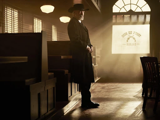 An older adult male with a grey beard wearing a dark coat and hat stands in a dimly lit room, illuminated by rays of light.