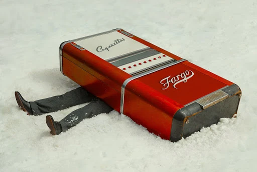 Red Fargo cigarette vending machine with a person's legs sticking out from underneath it in snow. Vintage-style visual and retro object.